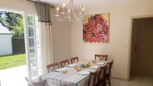 Un comedor con una mesa preparada para seis personas, rodeada de sillas de madera. Un candelabro cuelga sobre la mesa, y un cuadro grande con motivos florales adorna la pared.
