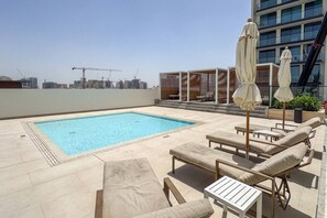 A rooftop pool area with lounge chairs and umbrellas, a clear view of the city skyline, and a modern building in the background.