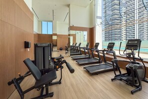 Une salle de sport équipée de tapis de course, d’appareils de musculation et dotée d’une grande fenêtre donnant sur un immeuble de grande hauteur.