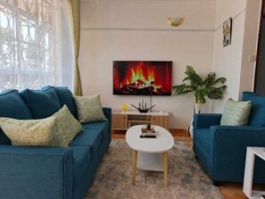 Een woonkamer met twee blauwe banken, een witte salontafel, een open haard met een schilderij erboven, en een potplant.
