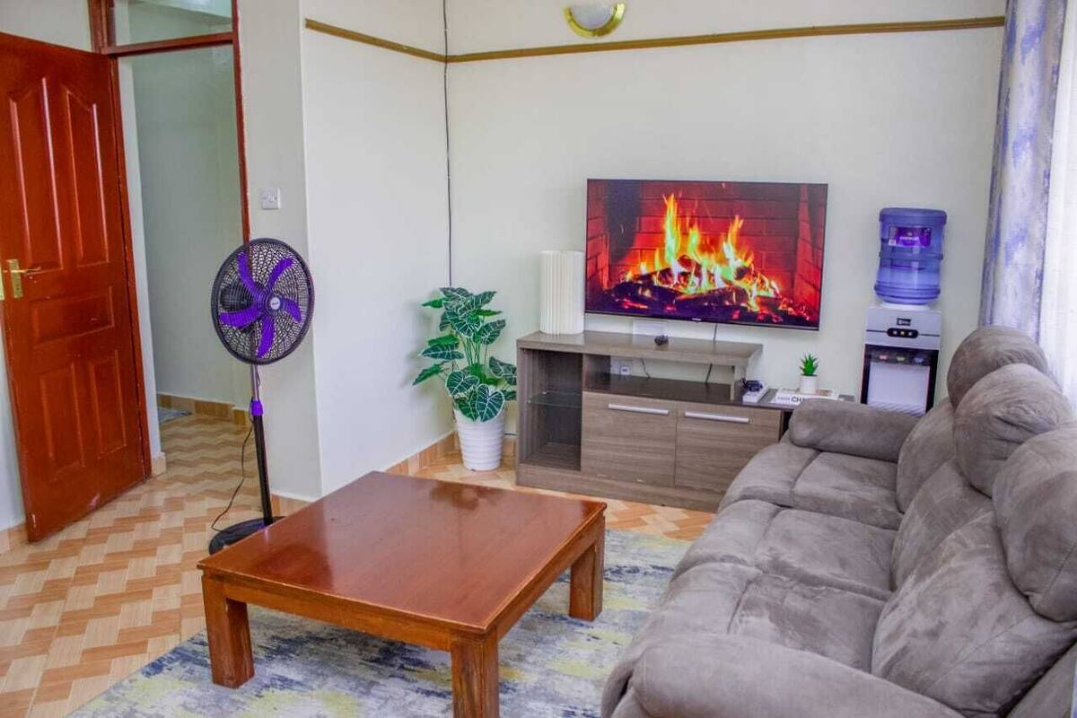 Un salon comprenant un canapé, une table basse en bois, un téléviseur diffusant une image de cheminée, un distributeur d’eau et un ventilateur.