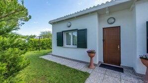 Une maison à l’extérieur blanc, avec des volets verts, une porte en bois et un petit jardin agrémenté d’une plante en pot.