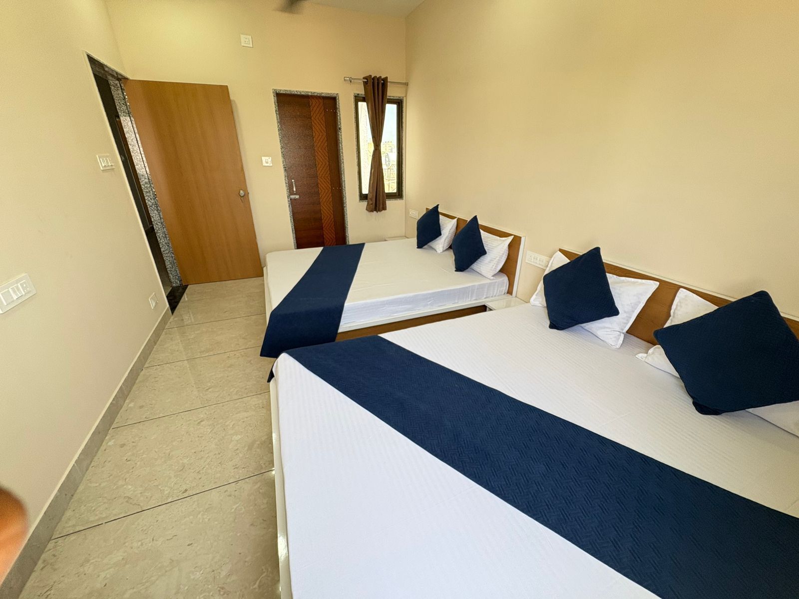 A hotel room with two beds, each with a blue bedspread and white pillows. There is a wooden door and a window with curtains.