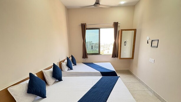 Habitación de hotel con dos camas, ventilador de techo, ventana con cortinas y vista a la ciudad.