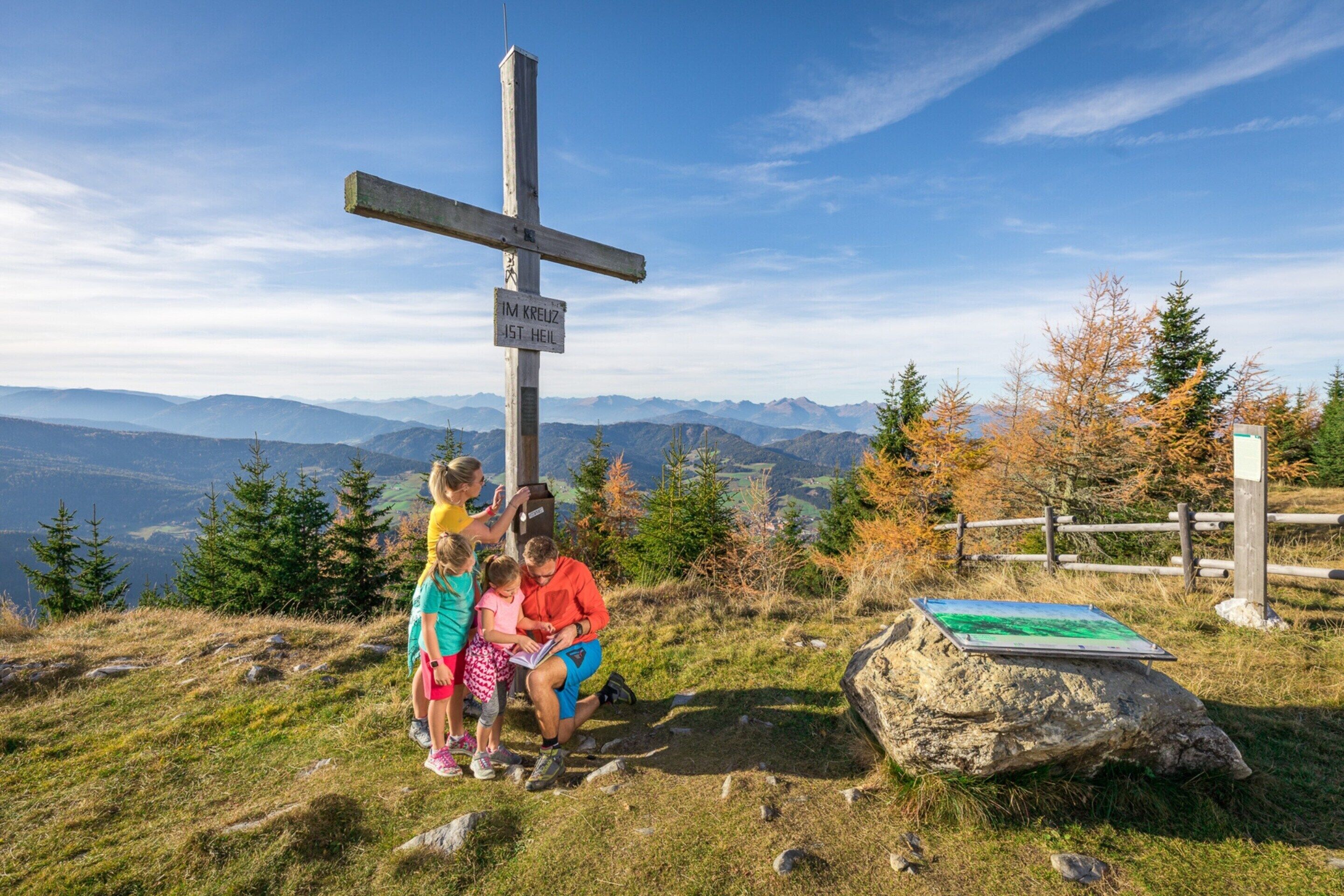 Una cruz de madera con un cartel que dice "IM KREUZ 1ST HEIL" en una zona montañosa con vistas a montañas distantes.