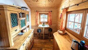 Interno di una baita di legno con un tavolo da pranzo, due sgabelli, un mobile con ante in vetro verde e una finestra con tende.