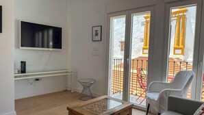 Ein Wohnzimmer mit einem Flachbildfernseher, einem Glastisch, einem weißen Sessel und einem Balkon mit Gartenmöbeln.