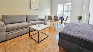 Una sala de estar moderna con un sofá gris, una mesa de centro blanca y un reposapiés gris. Hay una zona de comedor con sillas y una mesa, y un ventanal que proporciona luz natural.