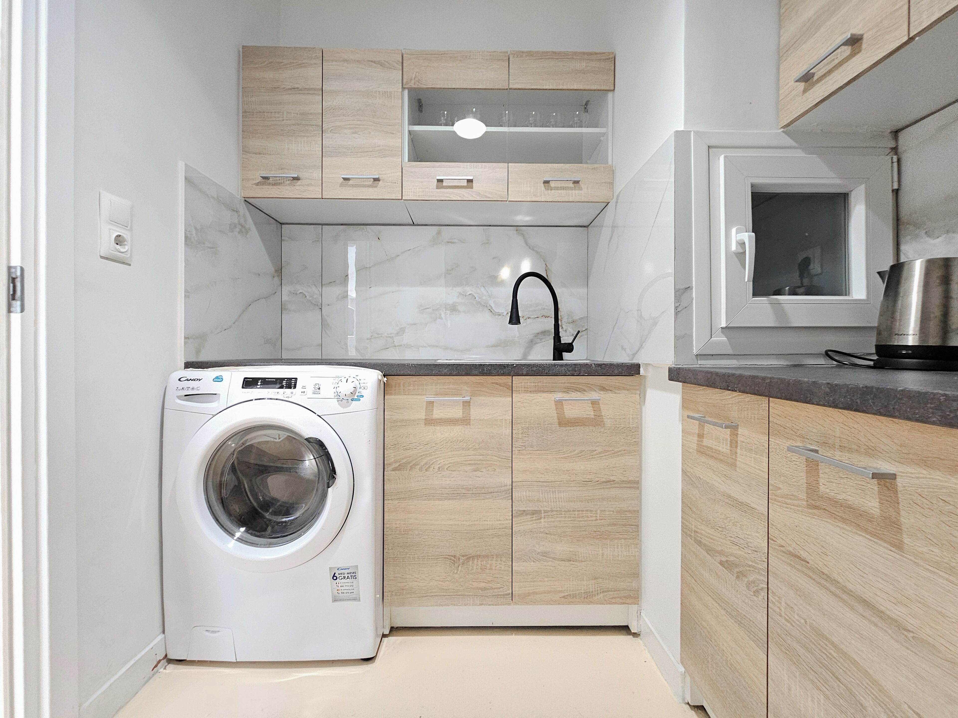 A compact kitchen with a built-in washer, wooden cabinets, a marble backsplash, and a stainless steel pot on the counter.