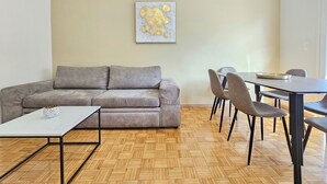 Una sala de estar moderna con un sofá gris, una mesa de centro blanca y una mesa de comedor con sillas. Hay un cuadro en la pared y un suelo de madera con patrón de espiga.