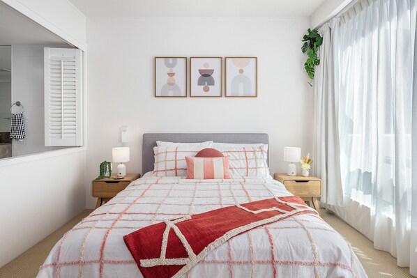 A bedroom with a bed, two bedside tables, a window with curtains, and three wall art pieces.