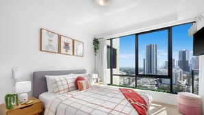 A modern bedroom with a large window, a bed with a patterned bedspread, a bedside table with a lamp, and framed artwork on the wall. - Stunning unit-perfect location (Gold Coast)