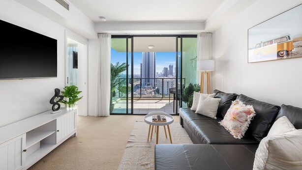 A modern living room with a large flat-screen TV, a sofa, a coffee table, and a balcony with a city view. - Stunning unit-perfect location (Gold Coast)