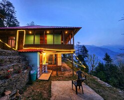 Una casa de madera de dos pisos con balcón, paredes de piedra y vista a las montañas.