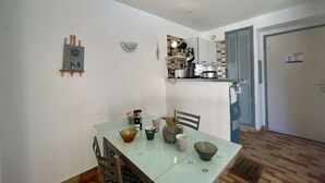 A small dining area with a table set for two, a kitchen in the background, and a door to the right. - 2-Room Apartment - Port Center - Cap d'Agde (Cap d'Agde)