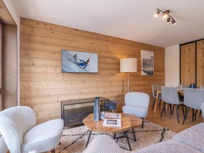 A modern living room with a wooden wall, a fireplace, a dining area with a table and chairs, and a mounted painting. - New apartment in Alpe d'Huez, ski-in ski-out (Huez)