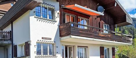 Chalet-style building with a balcony and sloped roof, surrounded by lush greenery and mountains.