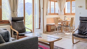 A cozy living room with a sofa, chairs, a coffee table, and a dining area with a table and chairs. Large windows with curtains allow natural light and views of the outdoors.