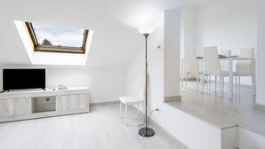 A modern living room with a TV, a white console table, a floor lamp, and a dining area with chairs and a table.