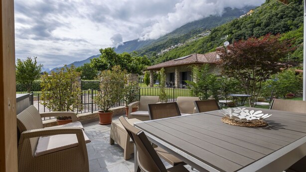 Eine Terrasse im Freien mit Tisch und Stühlen, umgeben von Pflanztöpfen und Blick auf einen Berg und ein Haus.