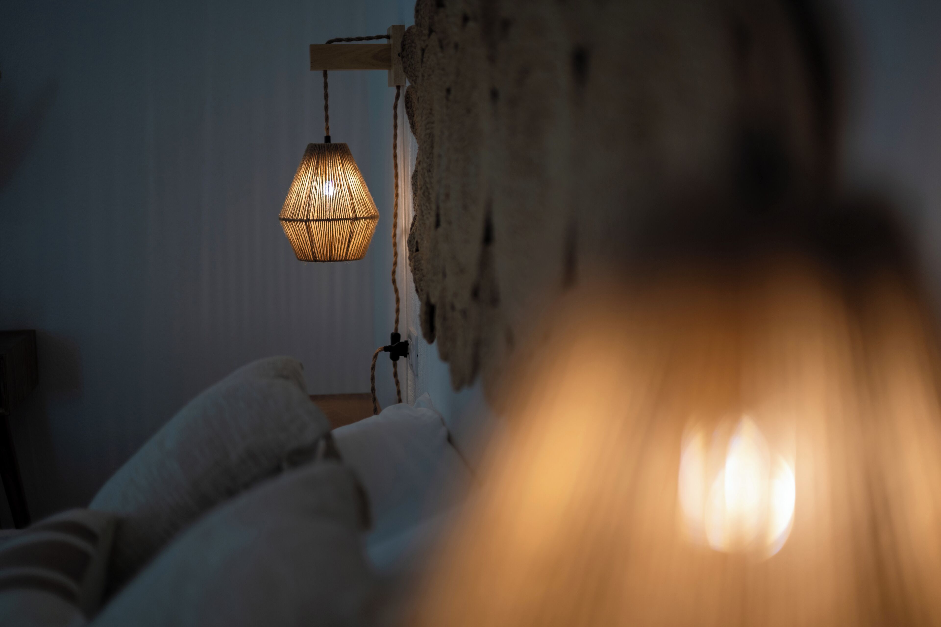 Une pièce faiblement éclairée, avec une lampe suspendue, un mur à texture et un lit recouvert de draps blancs.