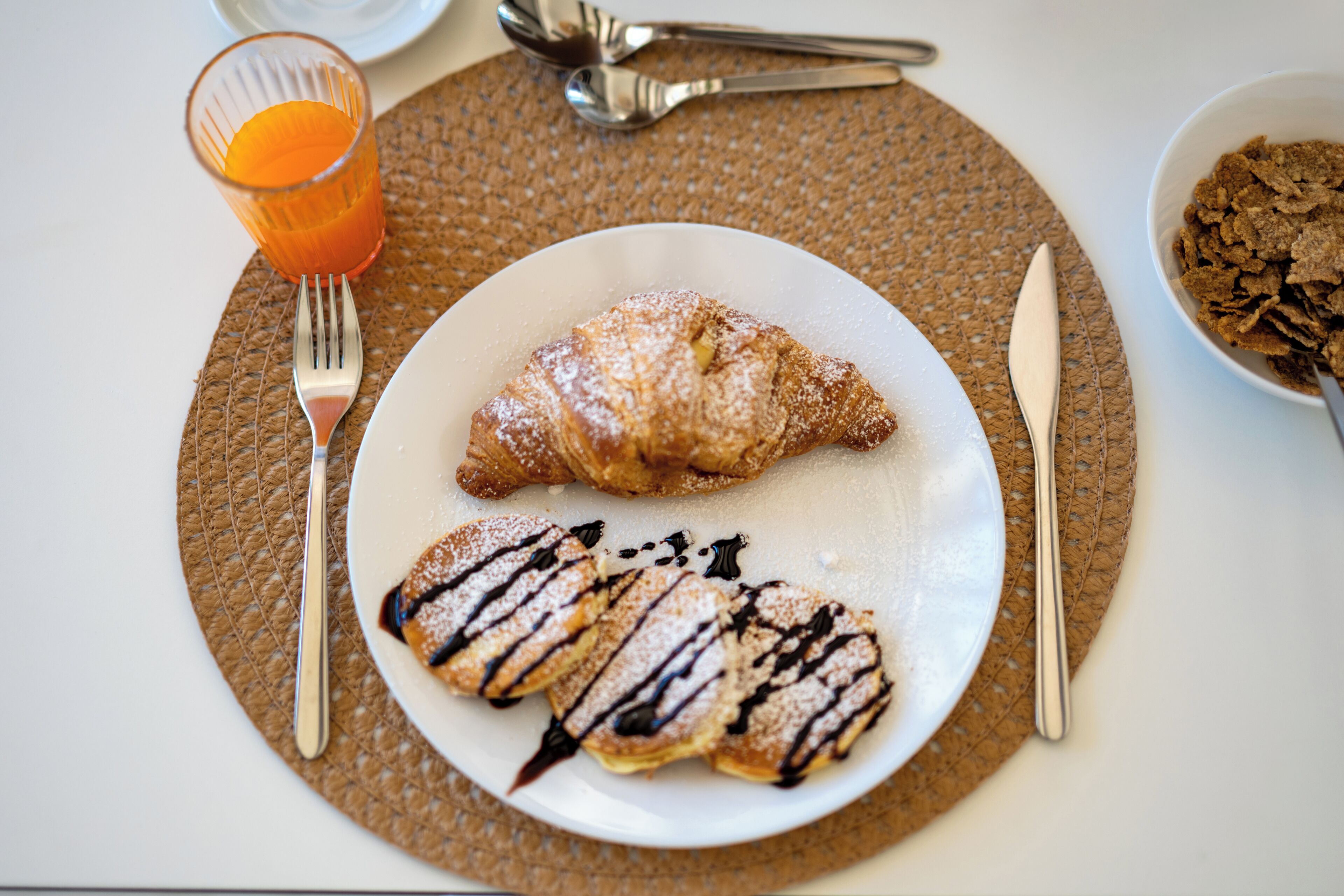 A breakfast plate with a croissant and waffles, a glass of orange juice, and a bowl of cereal.