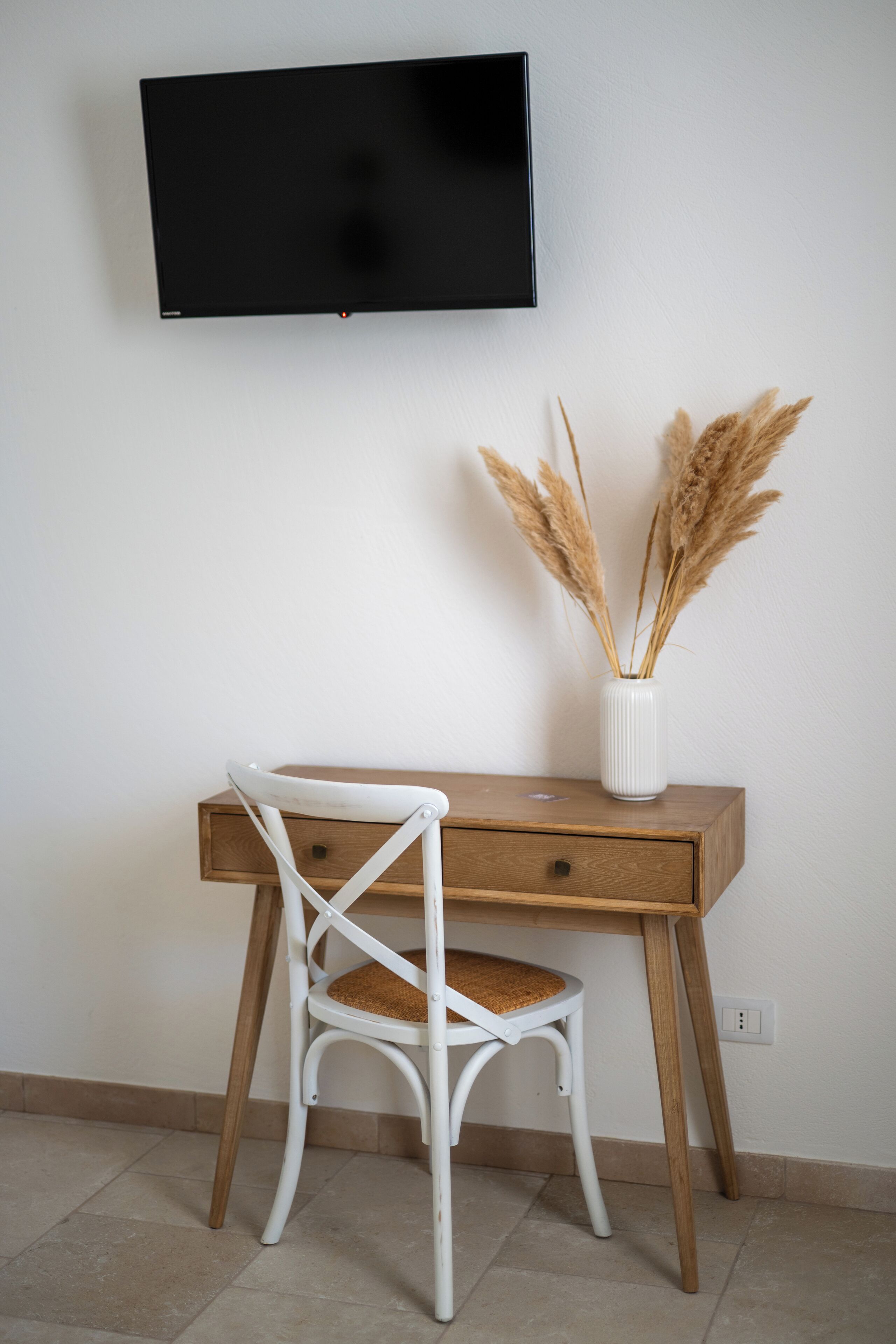 Una habitación con un escritorio de madera, una silla blanca, un televisor de pantalla plana montado en la pared y un jarrón con plumas de pluma de pampa seca.