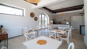A spacious dining area with white tables and chairs, a kitchen in the background, and a large window providing natural light. - Il Casale nel Parco (Ostuni)