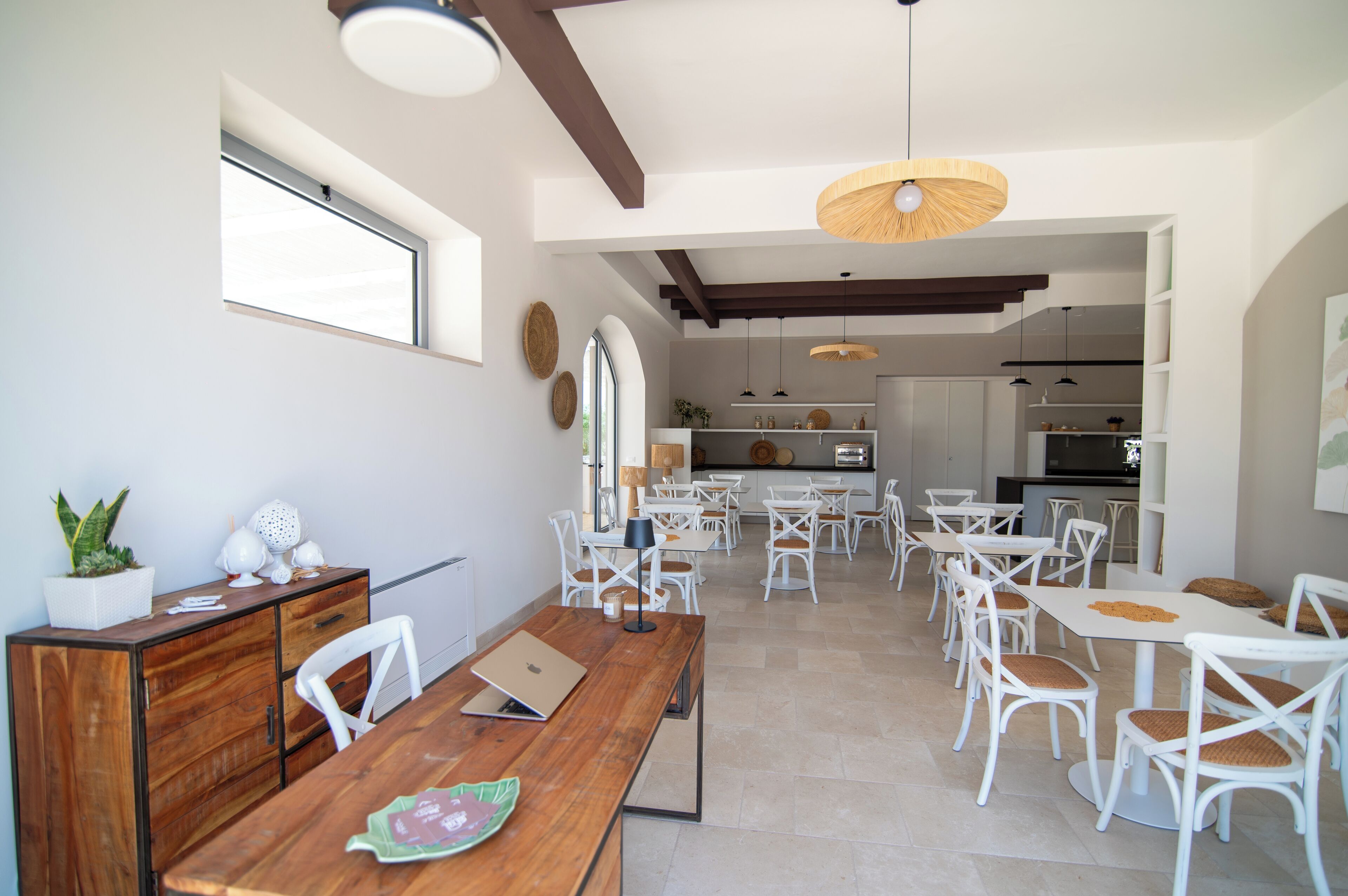 A spacious dining area with white chairs and tables, a wooden dining table, and a kitchen in the background.