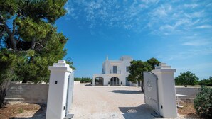 A white building with a symmetrical facade, a central entrance, and a gravel driveway leading to a gated entrance. - Il Casale nel Parco (Ostuni)