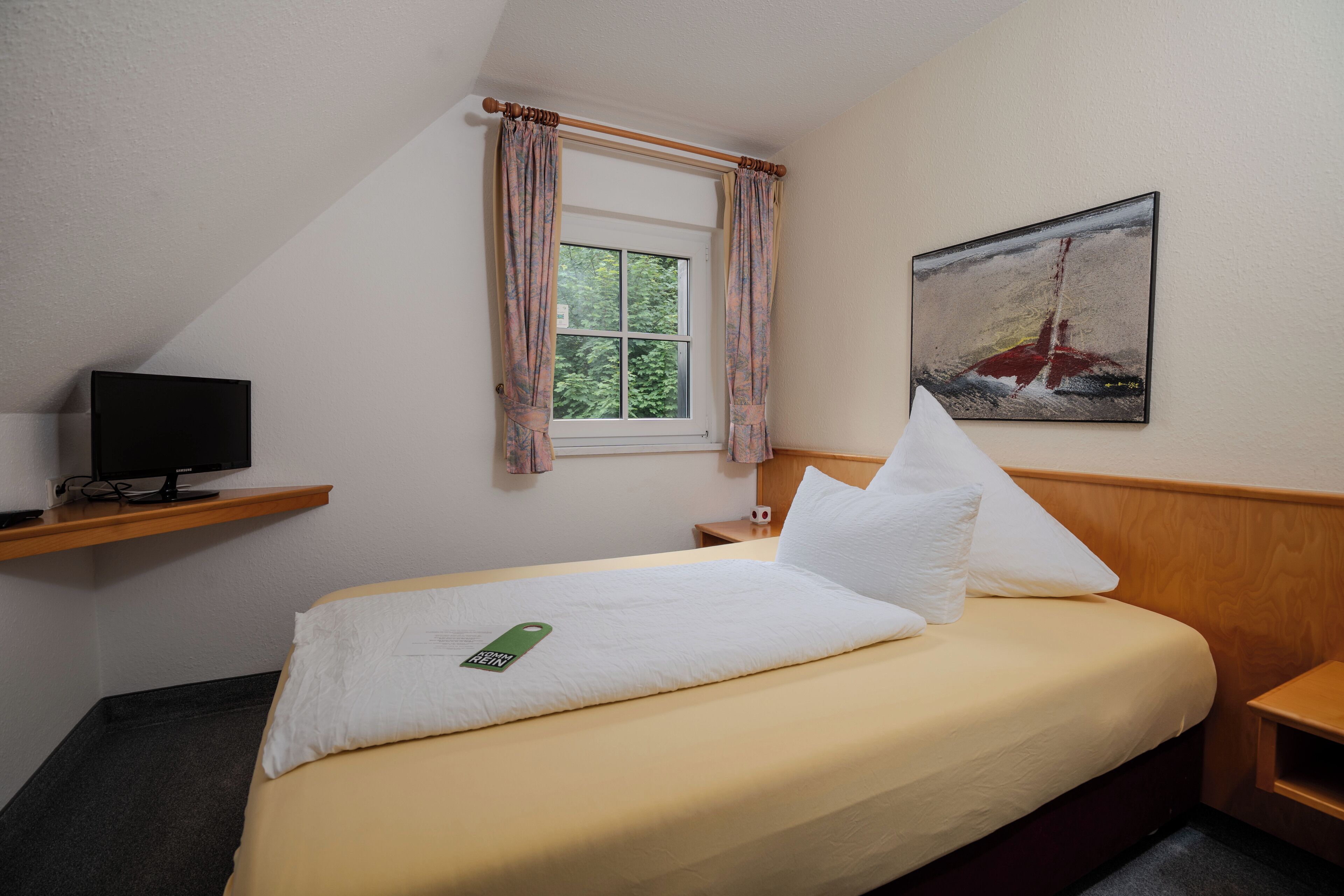 Een hotelkamer met een bed, een televisie op een plank, een raam met gordijnen, en een schilderij aan de muur.