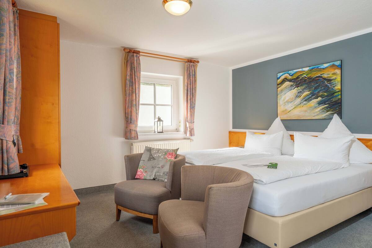 Habitación de hotel con cama, dos sillones, escritorio, ventana con cortinas, radiador y un cuadro en la pared.
