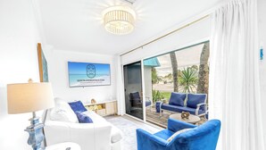A modern living room with a white sofa, blue armchair, and a remote control on a small table. A chandelier hangs from the ceiling, and a large window offers a view of the outdoors. - NEW LISTING! Village Breeze - Heated Pool, Private Patio, Village and Beach! (Siesta Key)