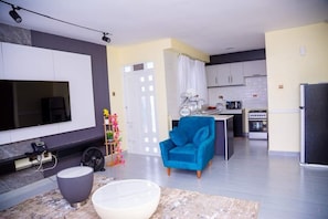 A modern living room with a flat-screen TV, a blue armchair, and a kitchen area with a stove and refrigerator.