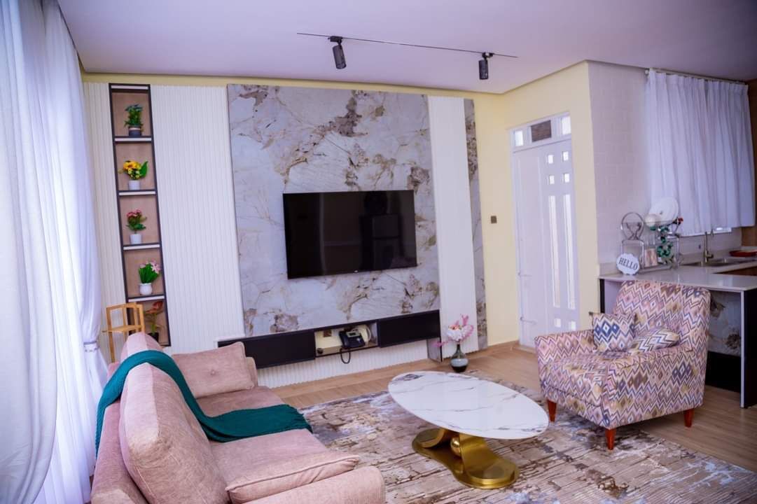 Un salon comprenant un canapé, un fauteuil à motifs, une cheminée en marbre, un téléviseur fixé au mur et une petite table avec un pied doré.