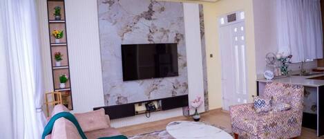 Un salon comprenant un canapé, un fauteuil à motifs, une cheminée en marbre, un téléviseur fixé au mur et une petite table avec un pied doré.