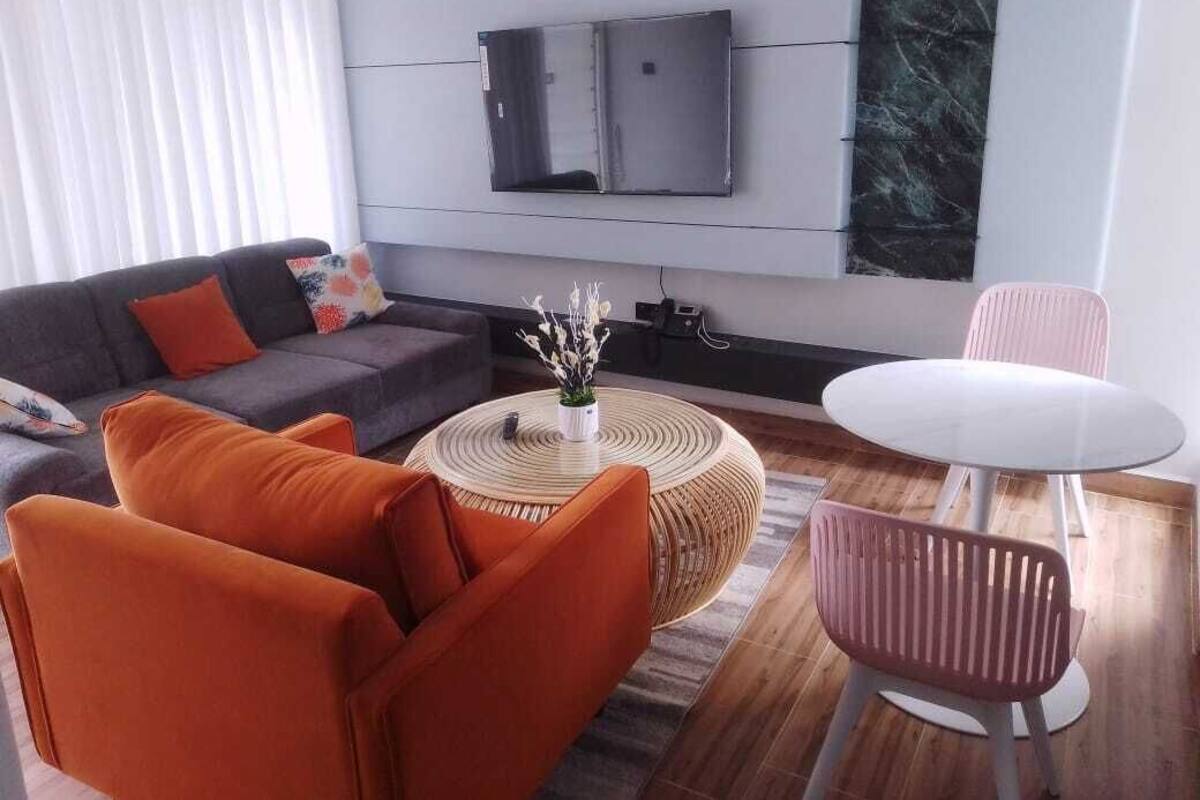Uma sala de estar moderna com um sofá cinzento, poltronas laranja, uma mesa de centro redonda e uma mesa de jantar branca.