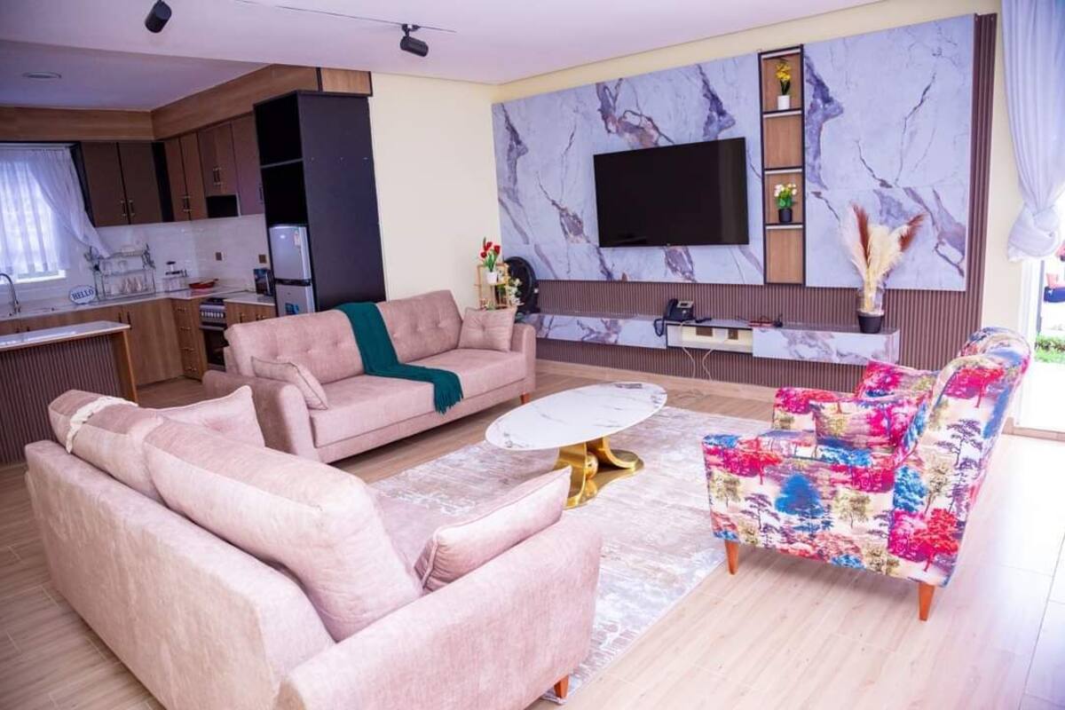 Sala de estar com sofá, mesa de centro e poltrona colorida. Há uma cozinha com armários e geladeira. Uma televisão de tela plana está fixada na parede.