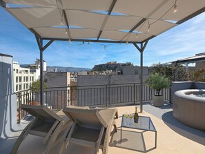 A rooftop terrace with lounge chairs, a table with glasses and a bottle, and a view of a historic cityscape. - The Acropolis Pathway (Athens)