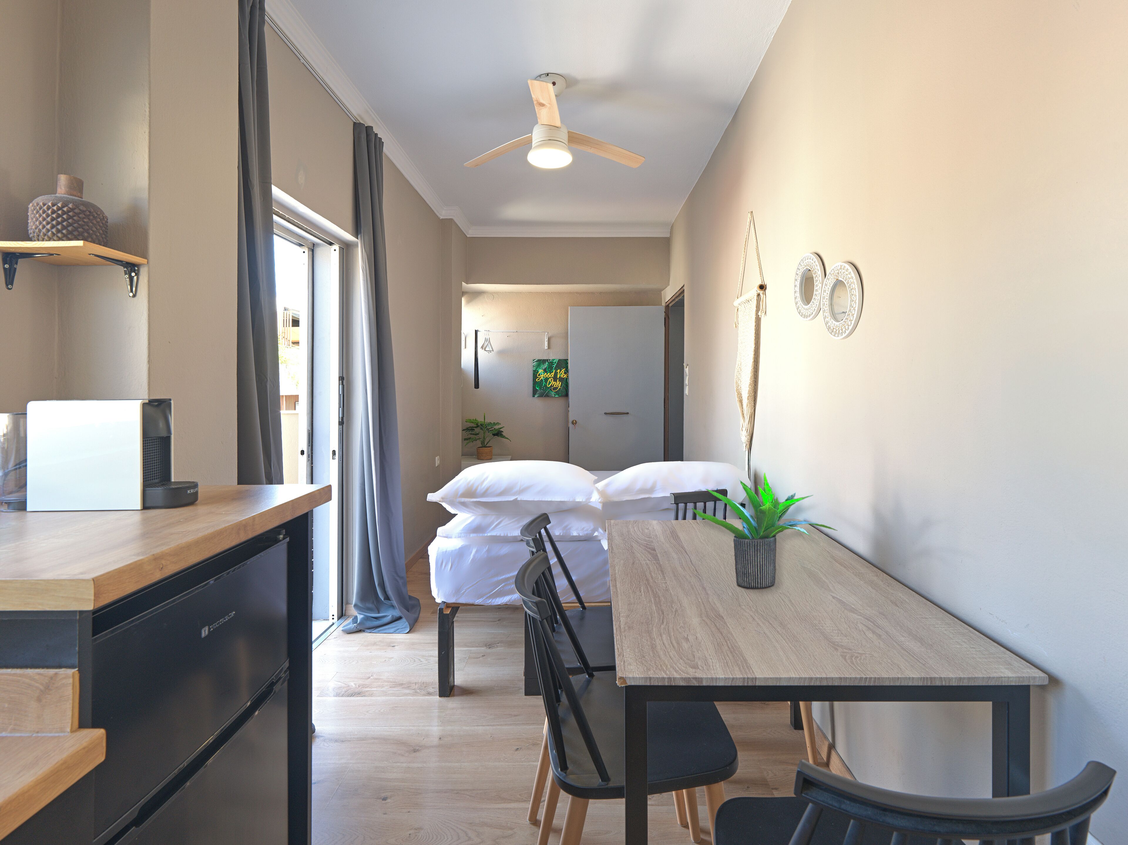 A modern, well-lit room with a wooden dining table, chairs, a ceiling fan, and a bed with white bedding.