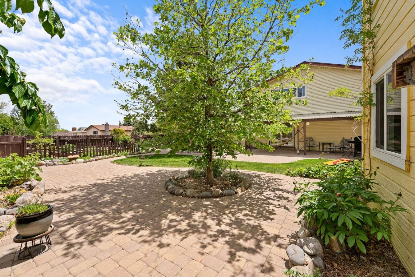 A paved courtyard with a central tree, a wooden fence, and a building with a covered patio area.