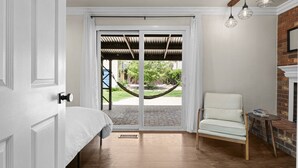 Un dormitorio con una cama, una silla, una mesita pequeña, una hamaca afuera y un muro de ladrillo.