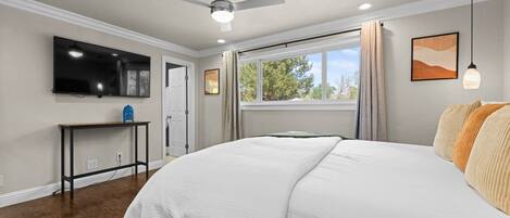 A bedroom with a bed, a TV, a ceiling fan, a window with curtains, and a desk.
