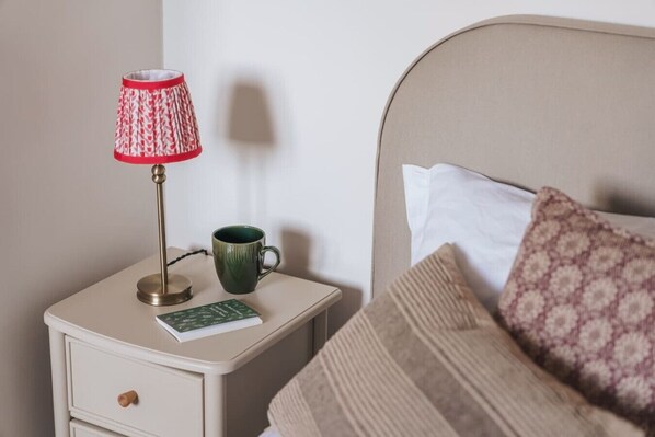 A neatly made bed with a patterned pillow, a bedside lamp with a red and white shade, a green mug, and a book on a bedside table.