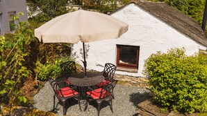 Une table ronde, quatre chaises et un parasol beige devant un bâtiment blanc doté de fondations en pierre et d’une fenêtre.