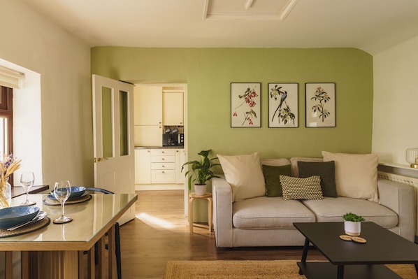 A living room with a sofa, a coffee table, and a dining area with a table and chairs. There is a kitchen in the background.