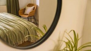Une pièce avec un lit recouvert d’une couverture à motifs verts, une chaise en bois avec un coussin et une plante en pot.