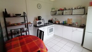 Une cuisine compacte avec des meubles blancs, une cuisinière, un four à micro-ondes et un évier. On y trouve une table à manger recouverte d’une nappe à carreaux rouges et oranges.