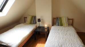 Un petit logement composé d’une seule pièce, comprenant deux lits simples, une table de chevet, une lampe et un puits de lumière.