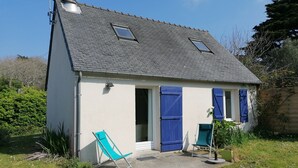 Une petite maison avec un toit en pente, deux fenêtres et une porte. Il y a deux chaises vertes à l’extérieur.